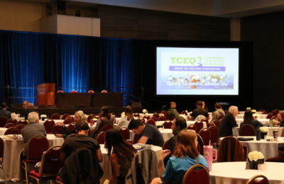 Crowd of people sitting at tables with a slide at the front of the room by the stage introducing the symposium